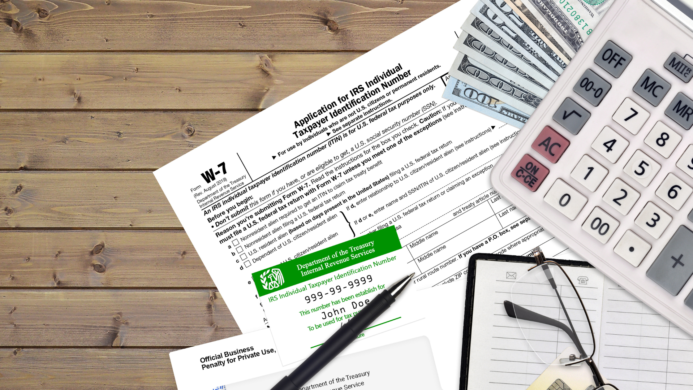 IRS W-7 tax form with calculator, money, pen, and glasses on a wooden desk.