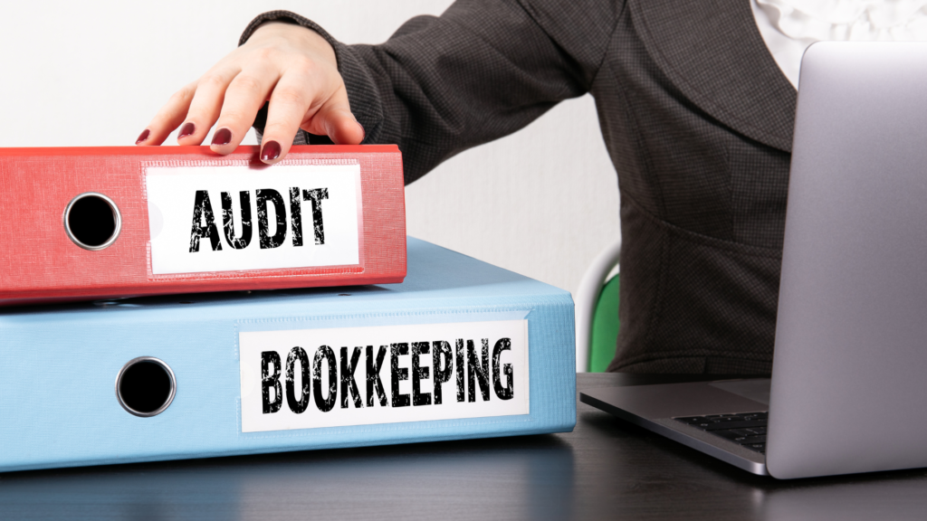 Close-up of red and blue binders labeled “Audit” and “Bookkeeping” beside a laptop.